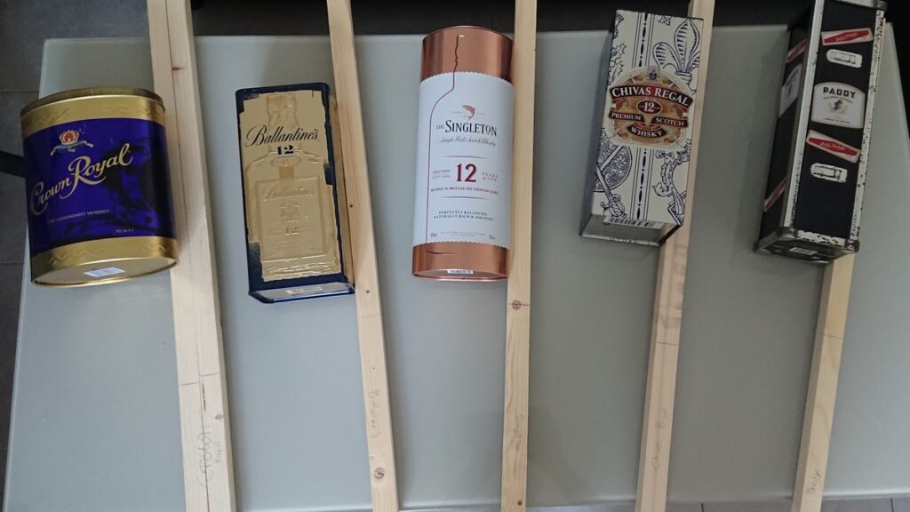 Cinq boites de bouteilles de whisky qui serviront de caisse de résonnance, à côté desquelles sont posés cinq tasseaux de sapin qui serviront de manche.